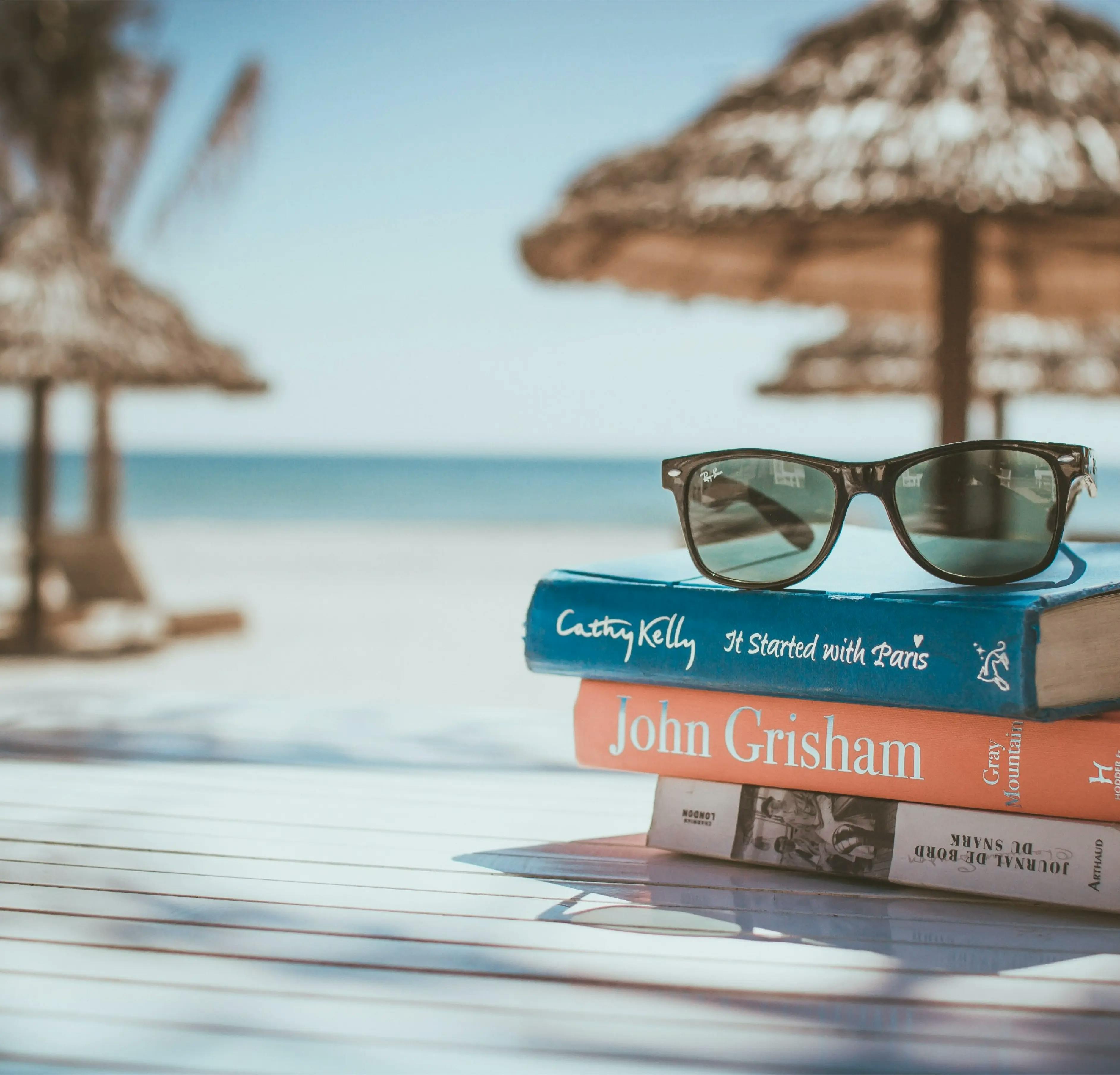 Blurred beach background with thatched umbrellas for shade. In the foreground, two books with sunglasses placed on top, in focus.