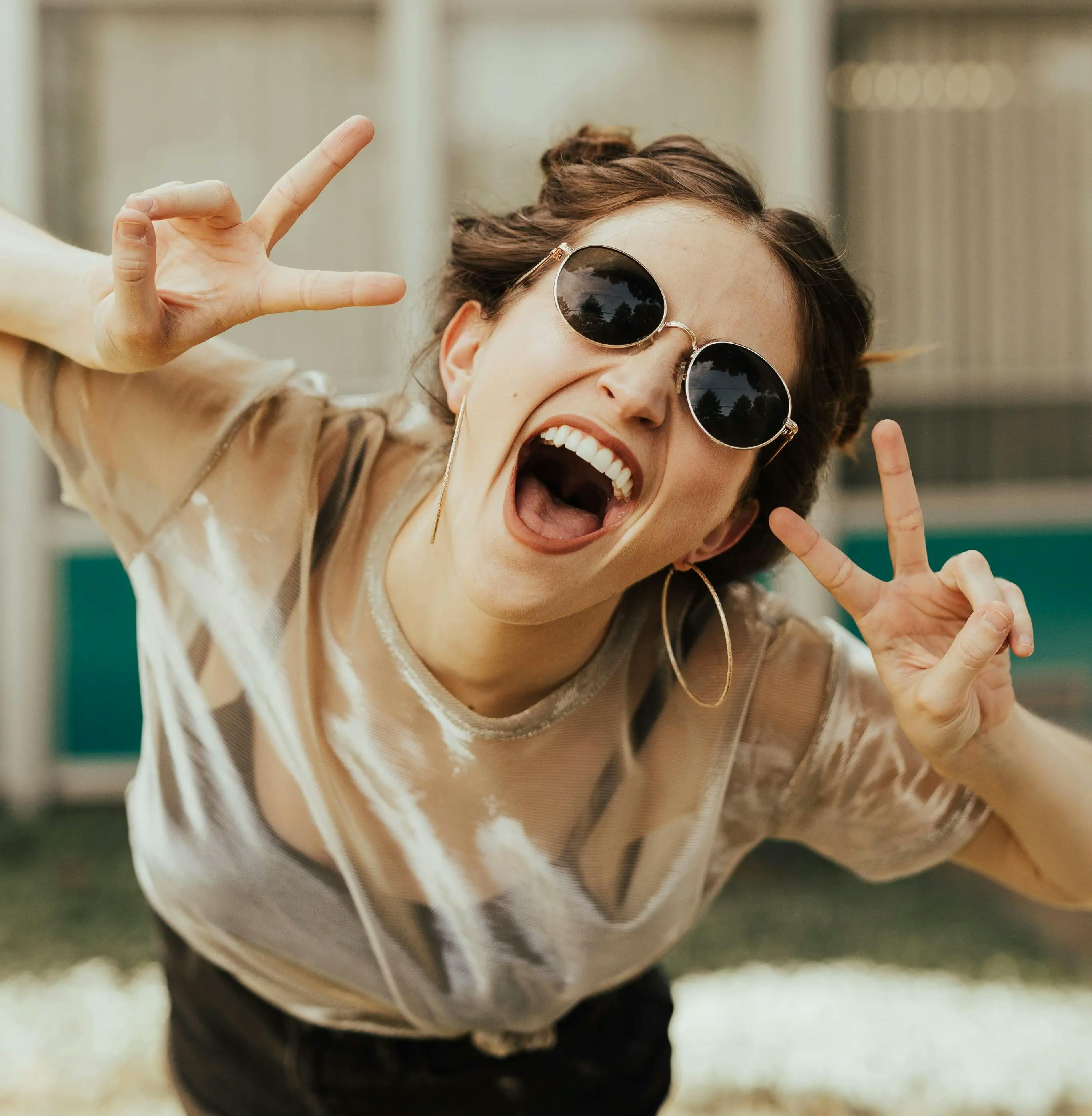 A smiling girl wearing sunglasses, making the peace symbol with both hands.